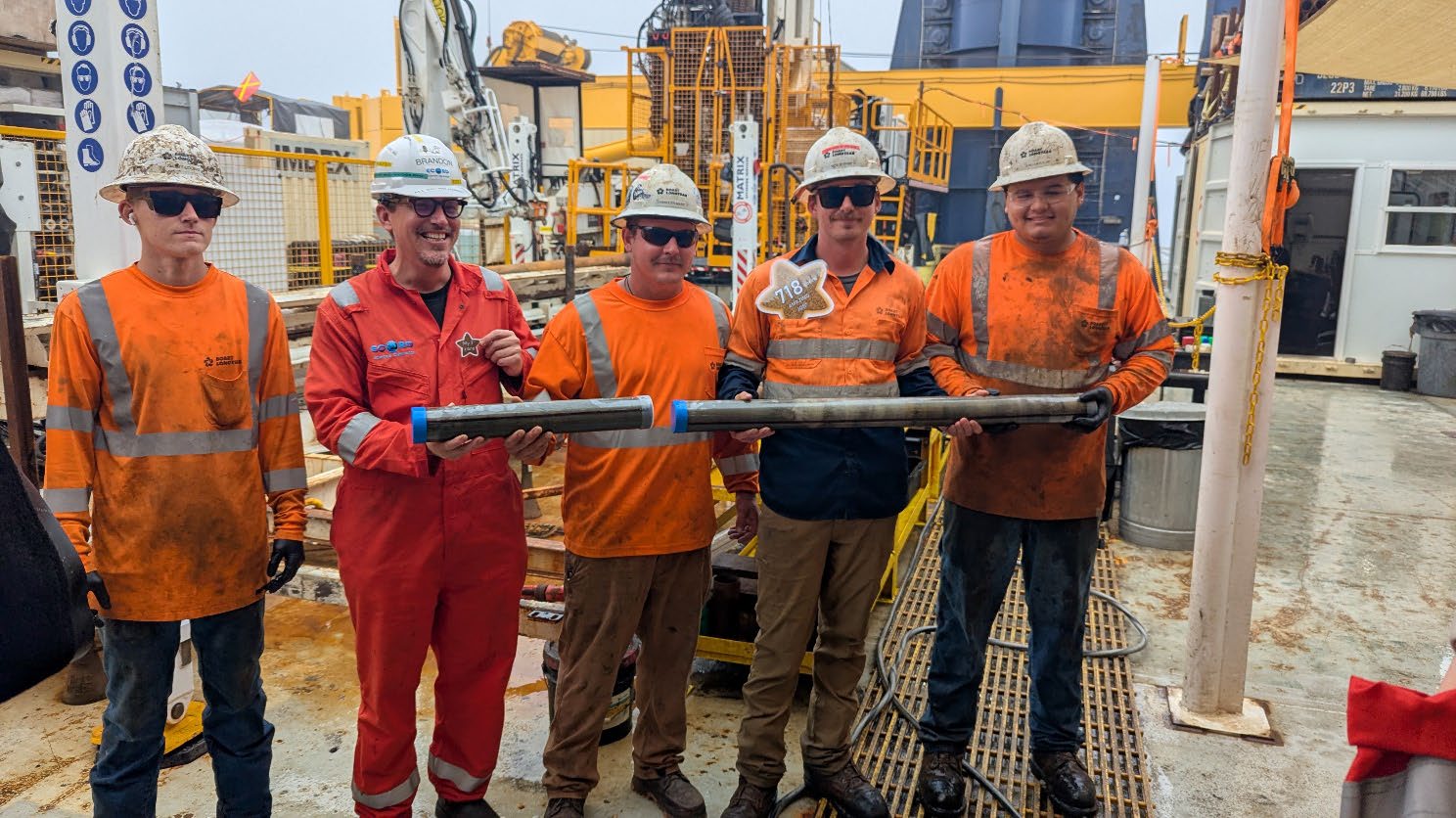 Four scientists in orange hi-vis and white hardhats stand on board a scientific research ship holding core material from the sea floor
